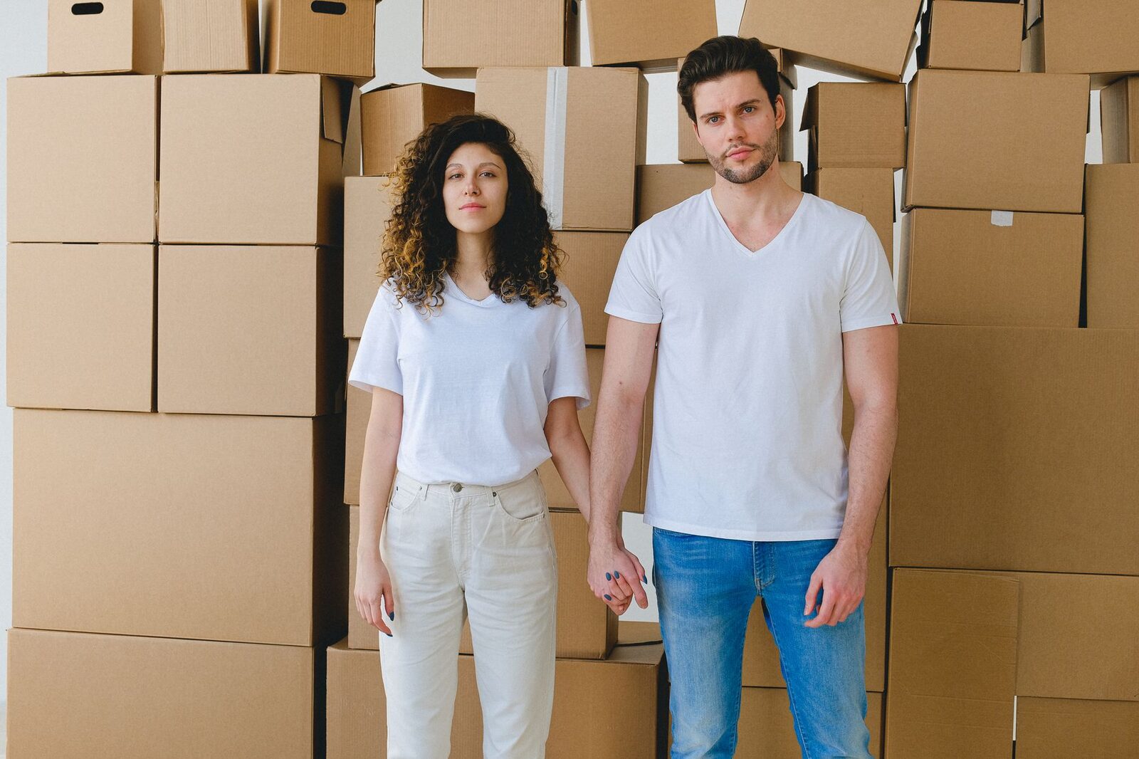 A young couple holding hands in front of a stack of cardboard boxes, possibly indicating moving or relocation.
